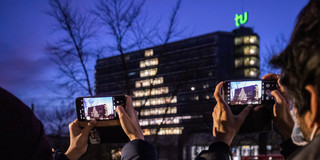 Two people taking a photo of the Math tower whose windows are illuminated in a christmas tree shape.