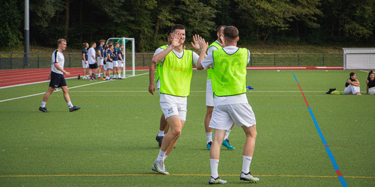 Photo: Two soccer players in white shirts and yellow vests high-five each other on the pitch.