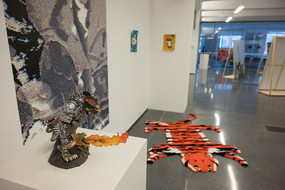 Im Vordergrund ist ein Tisch zu sehen, auf dem eine monsterhafte Figur steht. Im Hintergrund sieht man mehrere Kunstwerke, die in einem Raum stehen und zu denen auch ein auf dem Boden liegendes Objekt gehört, dass einem Tigerfell ähnelt. 