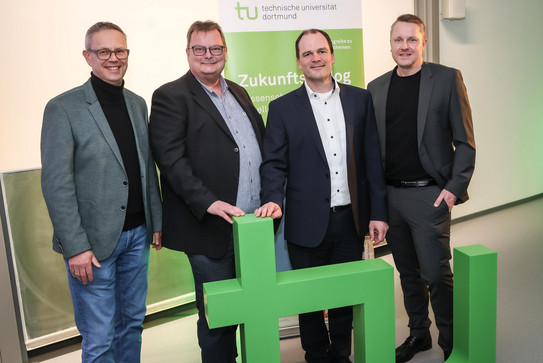 Das Bild zeigt vier männlich gelesene Personen, die hinter einem hüfthohen TU-Logo stehen. Im Hintergrund ist ein grün-weißer Aufsteller der TU Dortmund erkennbar.