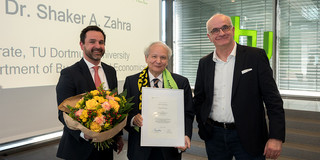 Das Bild zeigt die Professoren Strese, Zahra und Bayer zu sehen (v.l.) mit Blumen und einem Zertifikat. Dahinter ist eine Leinwand sowie eine Fensterfront zu sehen.