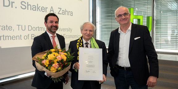 Das Bild zeigt die Professoren Strese, Zahra und Bayer zu sehen (v.l.) mit Blumen und einem Zertifikat. Dahinter ist eine Leinwand sowie eine Fensterfront zu sehen.