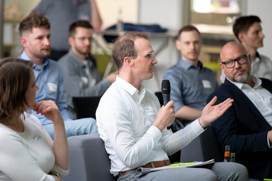 Foto: Ein Mann sitzt im Publikum und hält ein Mikrofon in der Hand. Er trägt ein weißes Hemd und Brille.