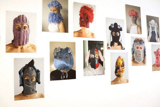 Auf dem Foto sieht man mehrere Fotografien, die an einer weißen Wand hängen und in der Ausstellung gezeigt werden. Die Fotografien zeigen Personen, die eine Gesichtsmaske aus unterschiedlichen Stoffen tragen.