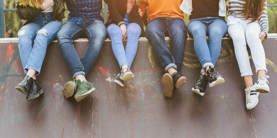 Several young people sit next to each other on a wall and dangle their feet. The faces of the people are not visible.