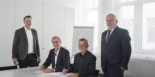 Gruppenfoto von vier Männern in Anzügen, zwei von ihnen sitzen an einem weißen Tisch und unterschreiben ein Dokument.