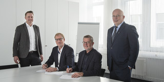 Group photo of four men in suits, two of them sitting at a white table signing a document.