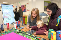 Das Bild zeigt einen Tisch mit ganz viele Utensilien. Auf dem Tisch liegt ein Tablet, dass von einem Kind bedient wird. Neben dem Kind stehen zwei erwachsene Frauen. Links im Bild sieht man ein Plakat, auf dem geschrieben steht, was Besucher*innen beim Hyle-C-Stand alles ausprobieren kann.