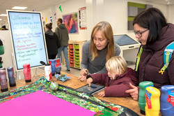 Das Bild zeigt einen Tisch mit ganz viele Utensilien. Auf dem Tisch liegt ein Tablet, dass von einem Kind bedient wird. Neben dem Kind stehen zwei erwachsene Frauen. Links im Bild sieht man ein Plakat, auf dem geschrieben steht, was Besucher*innen beim Hyle-C-Stand alles ausprobieren kann.