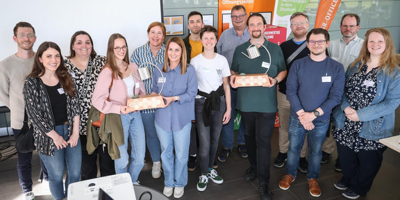 Gruppenfoto von 14 Personen: Einige Personen halten einen Preis in der Hand. Prof. Gerhard Schembecker von der TU Dortmund und Svenja Stepper, Kanzlerin der FH Dortmund, überreichen den Impact Cup an die Preisträger*innen.