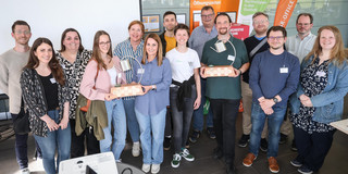 Gruppenfoto von 14 Personen: Einige Personen halten einen Preis in der Hand. Prof. Gerhard Schembecker von der TU Dortmund und Svenja Stepper, Kanzlerin der FH Dortmund, überreichen den Impact Cup an die Preisträger*innen.