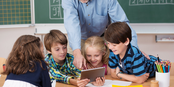 Four elementary school children are bent over a school desk. The teacher explains and shows them something on the tablet.