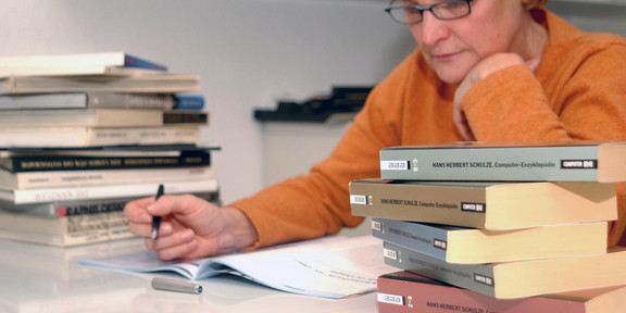 Eine Seniorin lernt umgeben von Bücherstapeln an einem Tisch