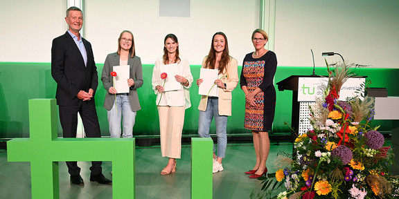Three young female students stand between a man and a woman. All three are holding certificates in their hands, with a large TU logo on the floor in the foreground.