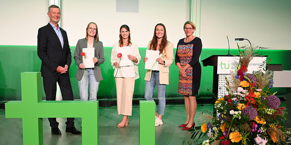 Drei junge Studentinnen stehen zwischen einem Mann und einer Frau. Alle drei halten Urkunden in den Händen, im Vordergrund steht ein großes TU-Logo auf dem Boden.