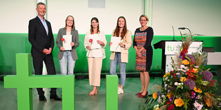 Three young female students stand between a man and a woman. All three are holding certificates in their hands, with a large TU logo on the floor in the foreground.