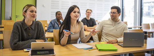 Studierende in einem Seminarraum in zwei Reihen. In der vorderen Reihe gestikuliert mittig eine Studentin. Die anderen Studierenden blicken in ihre Richtung.