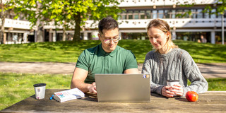 Zwei Studierende sitzen draußen an einem Tisch vor der Emil-Figge-Str. 50 und schauen gemeinsam auf den Bildschirm eines Laptops, der vor ihnen auf dem Tisch steht.