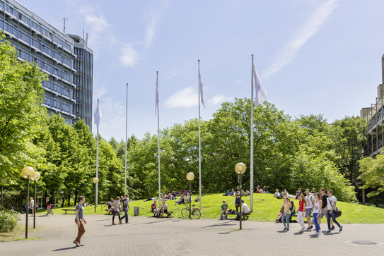 Gehisste TU Fahnen neben dem Mathetower. Blauer Himmel, grüne Umgebung im Sommer. Studenten laufen über den Campus.