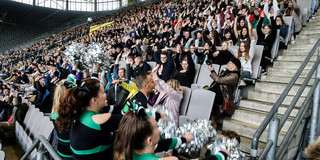 Die neuen Studierenden der TU Dortmund machen eine La-Ola-Welle auf einer gefüllten Tribüne im Fußballstadion.