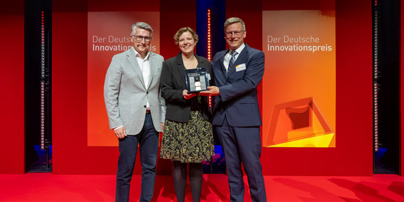 Foto: Drei Personen stehen auf einer Bühne mit einem Preis vor einer Wand mit der Aufschrift „Der Deutsche Innovationspreis“.