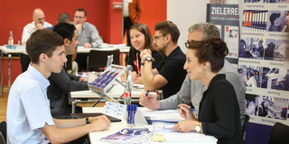 Karrieremesse Eine Beratungssituation. Zwei Personen sitzen an einem Tisch einer dritten gegenüber. Sie unterhalten sich.