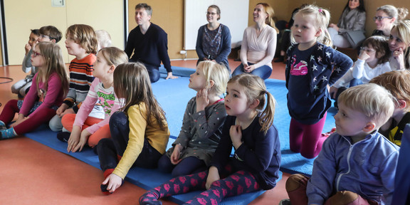 Eine Gruppe Kinder sitzt auf Matten auf dem Boden und schaut gespannt in eine Richtung. Im Hintergrund sitzen Erwachsene.