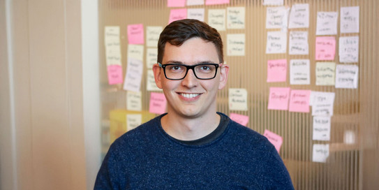 Porträtfoto von Prof. Sven Mayer: Prof. Mayer trägt eine Brille und einen blauen Pullover und steht vor einer braunen Wand mit bunten Post-Its.