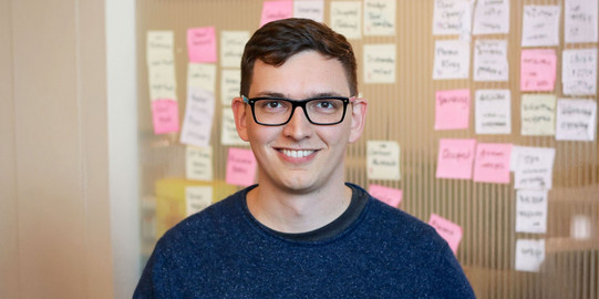 Porträtfoto von Prof. Sven Mayer: Prof. Mayer trägt eine Brille und einen blauen Pullover und steht vor einer braunen Wand mit bunten Post-Its.