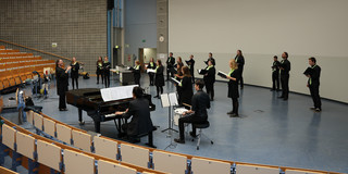 Ein Chor samt Dirigentin steht in einem Hörsaal und singt, einige Chormitglieder spielen ein Instrument.