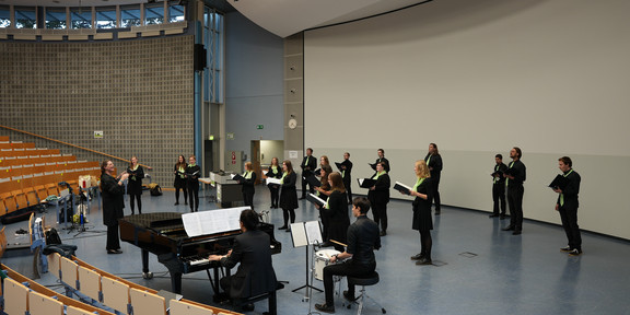 A choir including a conductor stands in an auditorium and sings, some choir members play an instrument.