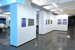 Das Bild zeigt weiße Ausstellungswände, an denen Leinwände hängen. Der Boden ist gemustert schwarz-grau glänzend.