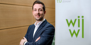 Simon Hensellek Ein Mann im Anzug posiert vor einem Poster mit dem Logo der wirtschaftswissenschaftlichen Fakultät.
