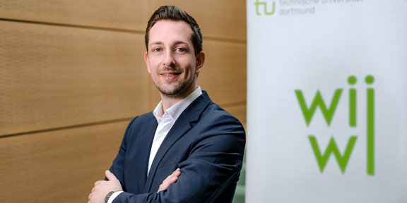 Simon Hensellek Ein Mann im Anzug posiert vor einem Poster mit dem Logo der wirtschaftswissenschaftlichen Fakultät.