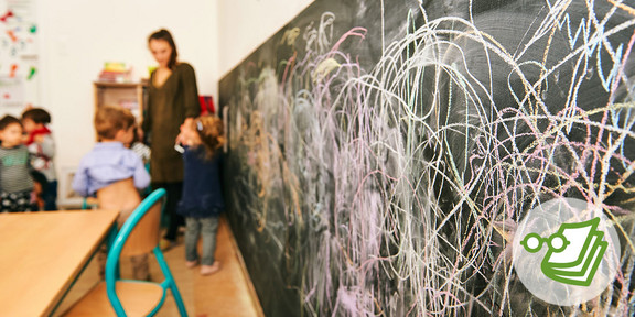 Eine vollgekrikkelte Tafel hinter der man unscharf Kinder und eine Lehrerin im Klassenzimmer sieht