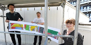 Drei Personen stehen auf einem Gerüst und halten eine Graphik, die die zukünftige Wandmalerei zeigt.