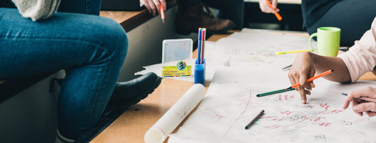 Brainstorming Vier Menschen sitzen an einem Tisch, die Gesichter sind nicht im Bild. Sie halten Stifte in der Hand und sehen nachdenkend aus. Eine Hand schreibt etwas auf das Plakat auf dem Tisch, welches sehr unstrukturiert gestaltet ist. Auf dem Tisch sind noch mehr Stifte und eine Tasse. Auf der Sitzbank erkennt man einen geöffneten Laptop und einen Rucksack im Hintergrund.