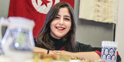 Eine internationale Studierende lächelt in die Kamera. Im Hintergrund hängt eine Tunesienflagge.