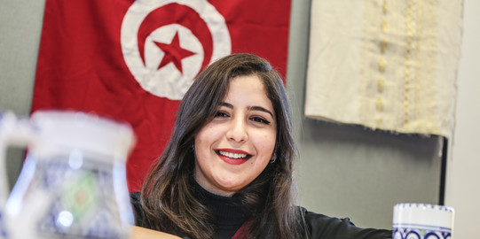 A young woman with brown hair sitting at a table and smiling. The flag of Tunisia can be seen on the wall behind her.