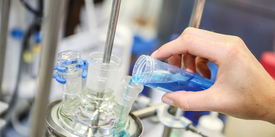 Eine Hand füllt eine türkisblaue Flüssigkeit in ein anderes Glasgefäß im Labor.