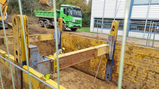 Hinter einem Bauzaun ist ein langer rechteckiger Aushub, in dem ein Bauarbeiter mit gelber Schutzjacke und Helm arbeitet. Gelbe Metallstützen stützen die Seiten des Aushubs ab. Auf der gegenüberliegenden Seite der Baustelle parkt ein dunkelgrüner LKW.