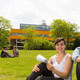 Zwei Studierende sitzen auf einer Wiese, im Hintergrund die Mensa