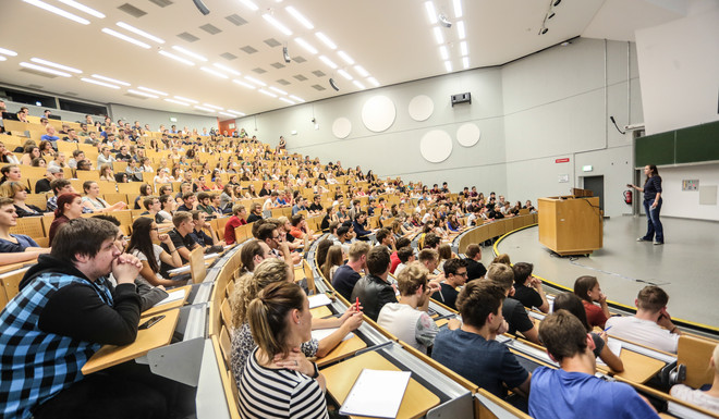Vorlesung im Hörsaal Viele Studierende sitzen bei einer Vorlesung im Hörsaal. Vorne steht eine Dozentin.