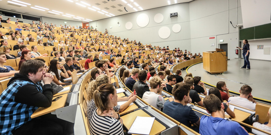 Vorlesung im Hörsaal Viele Studierende sitzen bei einer Vorlesung im Hörsaal. Vorne steht eine Dozentin.