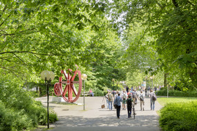 Studierende sind im Sommer auf dem Campus Nord, rote Zahnräder im Hintergrund.