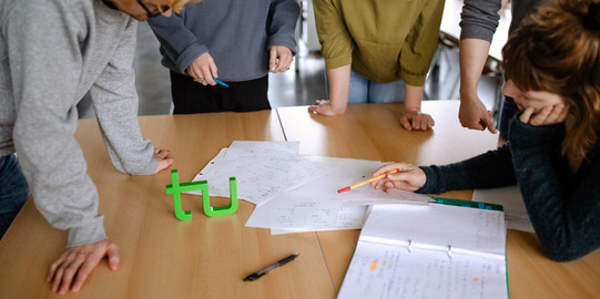 fünf Studierende stehen um einen Tisch und lesen ein Dokument.