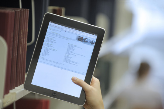 Eine Hand hält ein Tablet in der Universitätsbibliothek an einem Bücherregal.