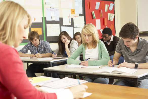 Schülerinnen und Schüler sitzen in einer Klasse und schreiben in Hefte. Vor ihnen sitzt eine Lehrerin an einem Pult.