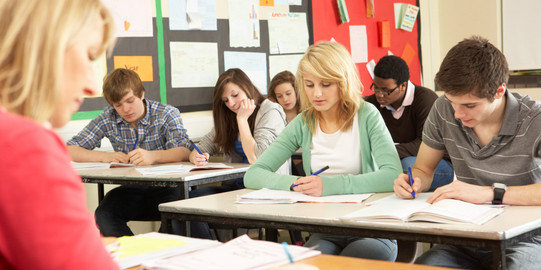 Unterricht im Klassenraum Schülerinnen und Schüler sitzen in einer Klasse und schreiben in Hefte, vor ihnen sitzt eine Lehrerin an einem Pult.