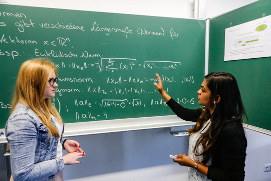 Zwei Personen stehen vor einer Tafel, auf die mit Kreide Formeln geschrieben sind. Die Person rechts zeigt mit dem Arm auf die Tafel und spricht. Die Person links sieht die Person rechts an.